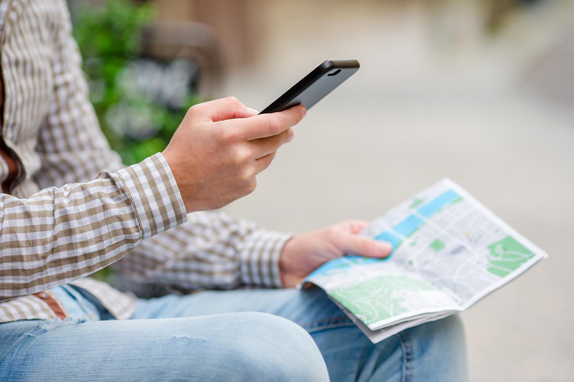 Closeup of male hands holding cellphone and city map outdoors on the street. Man using mobile