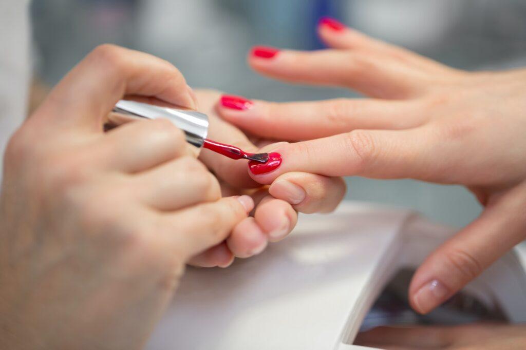 Woman nail master doing nails to a girl client at a beauty salon. A beautician applies red nail